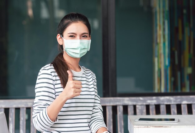 Distanciamiento social Mujer joven asiática que usa mascarilla médica y muestra los pulgares hacia arriba Nuevo concepto normal y de atención médica