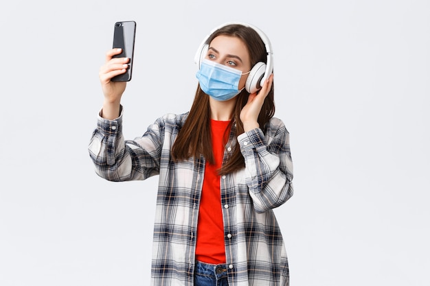 Foto gratuita distanciamiento social, ocio y estilo de vida en el brote de covid-19, concepto de coronavirus. mujer en auriculares y máscara médica escuchando música, tomando selfie en teléfono móvil con filtros.