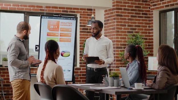 Diversas personas que presentan proyectos y estrategias de negocios en la oficina de reuniones. Compañeros de trabajo haciendo presentaciones financieras en el monitor frente a colegas, trabajando en desarrollo y crecimiento.