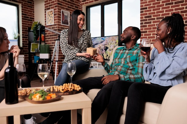 Diversas personas sonrientes disfrutando de la fiesta del vino en casa mientras intercambian regalos y beben vino. Jóvenes grupos multiétnicos de amigos que se unen mientras se dan regalos en casa en la sala de estar.