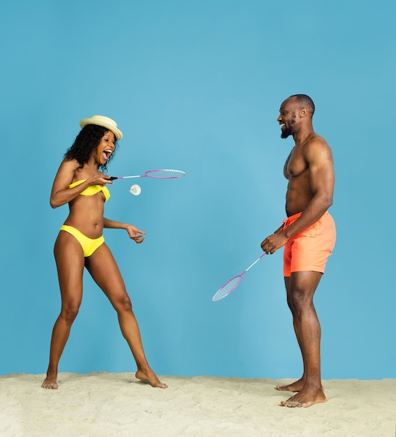 Diversión más loca. Feliz pareja joven afroamericana jugando bádminton sobre fondo azul de estudio. Concepto de emociones humanas, expresión facial, vacaciones de verano o fin de semana. Frío, verano, mar, océano.