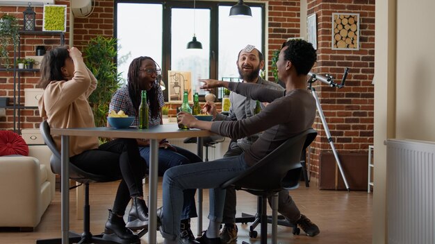 Diverso grupo de amigos divirtiéndose juntos con juegos de cartas, disfrutando de la actividad estratégica en la reunión. Gente moderna jugando juegos de mesa con bebidas alcohólicas y bocadillos en casa.