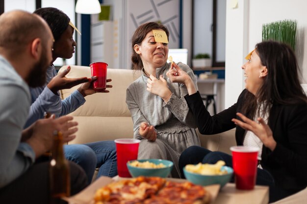 Diverso grupo de compañeros de trabajo jugando a adivinar quién con notas pegajosas en la frente, disfrutando de actividades divertidas con juegos de charadas. Gente adivinando pantomima en la celebración de bebidas después de las horas de trabajo.