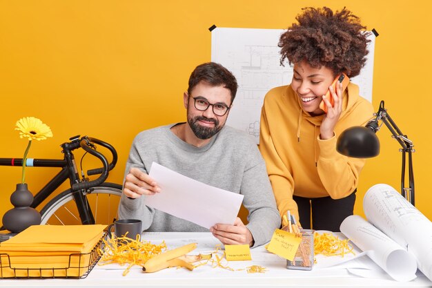 Diversos hombres y mujeres satisfechos se divierten en la oficina mientras preparan un proyecto creativo, colaboran para una tarea común en el espacio moderno, hacen papeleo, crean bocetos y planos. Dos arquitectos cerca del escritorio.