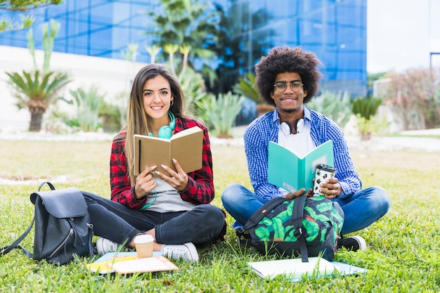 Diversos pares jovenes que sostienen los libros en la mano que se sienta en césped en el campus universitario