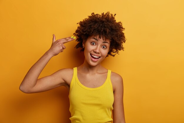 Divertida y alegre mujer de piel oscura se suicida, hace una pistola con los dedos, inclina la cabeza y dispara en la sien, hace el tonto y se aburre, viste ropa casual amarilla. Gente, estilo de vida, emociones