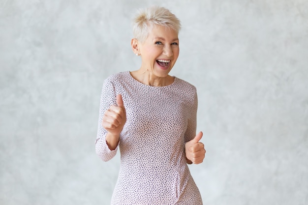 Divertida y alegre mujer rubia de mediana edad con elegante vestido lápiz bailando y mostrando los pulgares hacia arriba gesto como señal de aprobación, celebrando el éxito o un trato rentable, sonriendo ampliamente