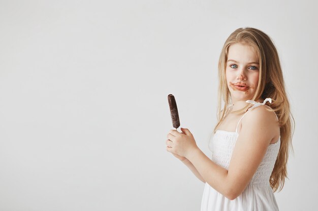 Divertida alegre niña con cabello claro en vestido blanco con cara sucia. El niño ya comió un helado y tendrá uno más.