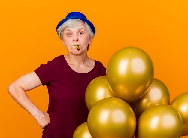 Divertida anciana con sombrero de fiesta se encuentra con globos de helio que soplan silbato aislado en la pared naranja con espacio de copia