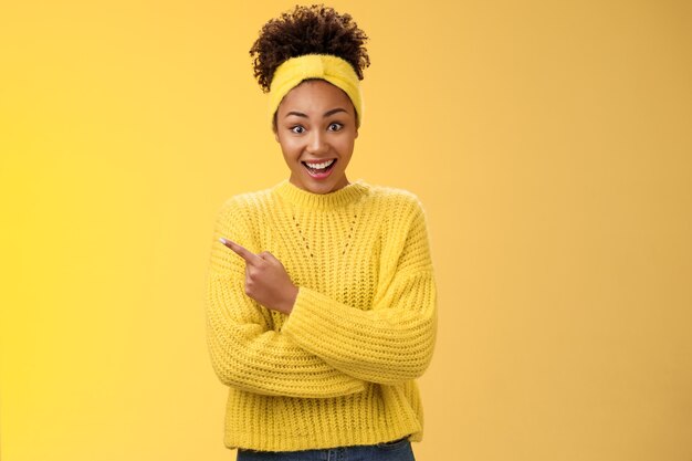 Divertida, carismática, sonriente, linda chica negra en suéter con diadema, abrir los ojos con la mandíbula caída, asombrado, escuchar acerca de un nuevo lugar interesante e interesante hablando amigo parado fondo amarillo apuntando a la izquierda asombrado.