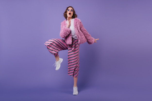 Divertida chica alegre europea en zapatillas blancas posando en la pared púrpura. Señorita elegante en pantalones a rayas bailando.