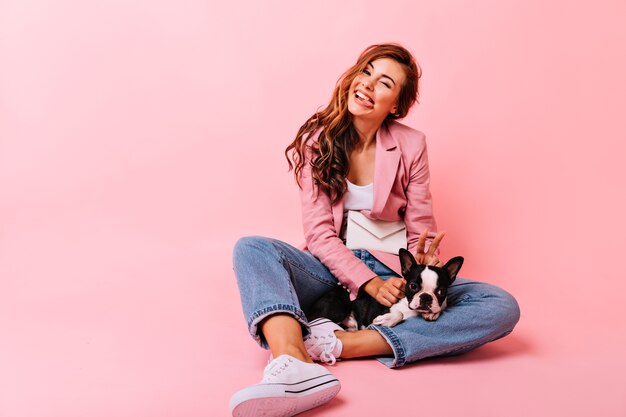 Divertida chica blanca con bulldog francés haciendo muecas. Retrato de interior de alegre mujer caucásica sentada en el suelo con cachorro.