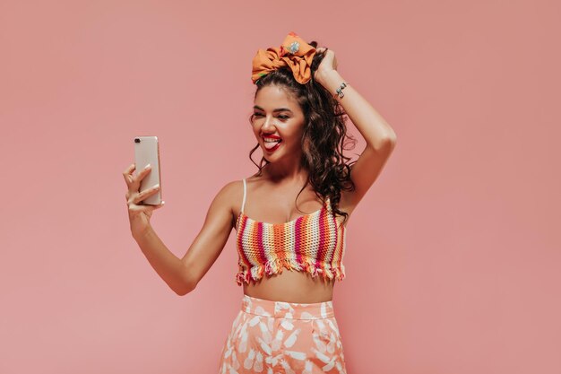 Divertida chica rizada con elegante diadema y accesorios en top colorido y pantalones estampados de piña hace foto sobre fondo rosa