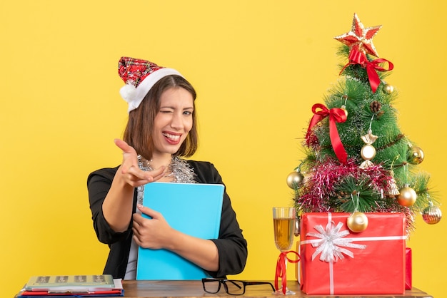 Divertida dama encantadora en traje con sombrero de santa claus y decoraciones de año nuevo con documento en la oficina en amarillo aislado