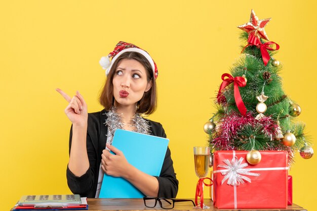 Divertida dama encantadora en traje con sombrero de santa claus y decoraciones de año nuevo con documento pensando profundamente en la oficina en amarillo aislado