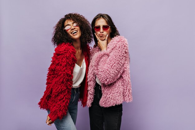 Divertida dama morena vestida con ropa mullida rosa y gafas de sol rojas posando con una chica fresca rizada en jeans y suéter rojo cálido