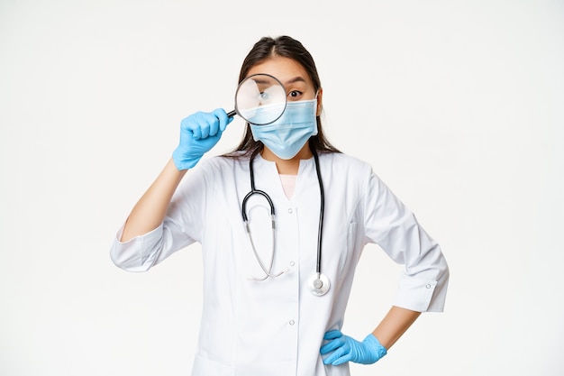 Divertida doctora asiática mira a través de una lupa al paciente, usa mascarilla médica y guantes de goma, fondo blanco.