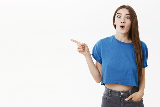 Divertida y emocionada morena atractiva entusiasta en elegante camiseta azul jadeando labios plegables de asombro apuntando a la izquierda