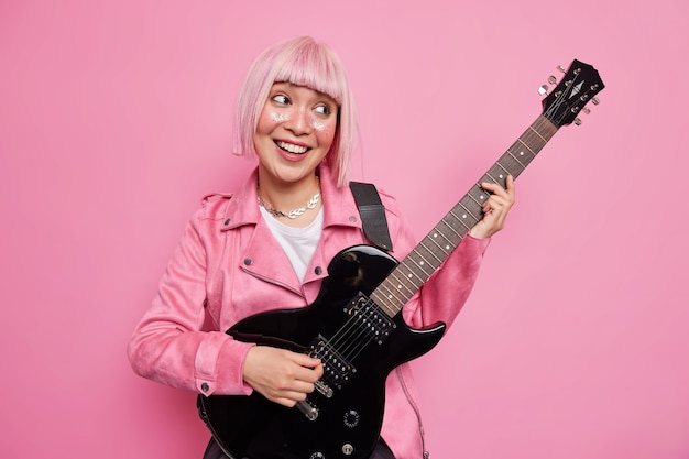 Foto gratuita divertida estrella de rock de pelo rosa alegre toca la guitarra eléctrica siendo parte de la banda vestida con chaqueta lista para improvisar en el escenario realiza nuevas poses de canciones en el interior se divierte. concepto de pasatiempo de entretenimiento musical