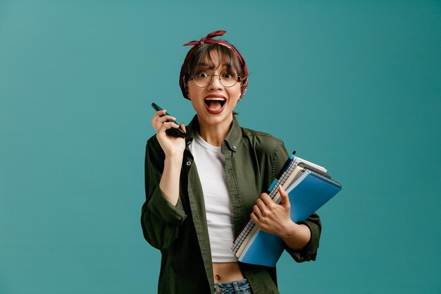 Divertida joven estudiante con gafas de bandana sosteniendo grandes blocs de notas con bolígrafo y teléfono móvil cerca de la oreja mirando la cámara aislada en el fondo azul