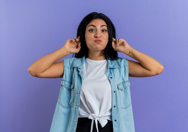 Foto gratuita divertida joven mujer caucásica soplando las mejillas y tomados de la mano detrás de las orejas aislado sobre fondo púrpura con espacio de copia