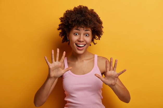 Foto gratuita divertida joven rizada levanta las palmas y tiene un estado de ánimo juguetón, tonta, sonríe feliz, usa una camiseta informal, expresa buenas emociones y sentimientos sinceros, aislado en la pared amarilla