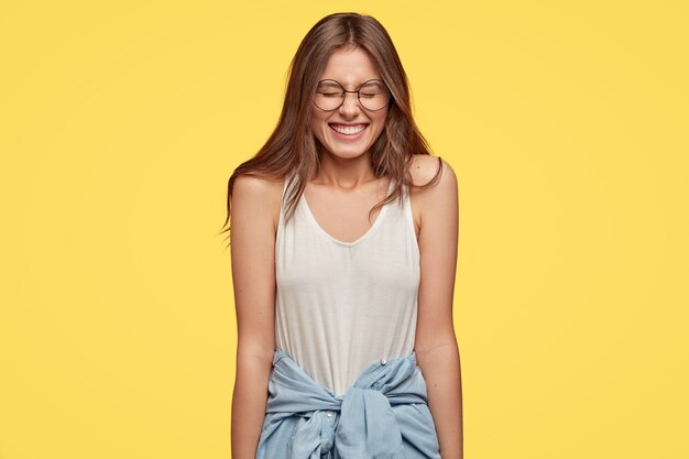 Divertida morena joven con gafas posando contra la pared amarilla