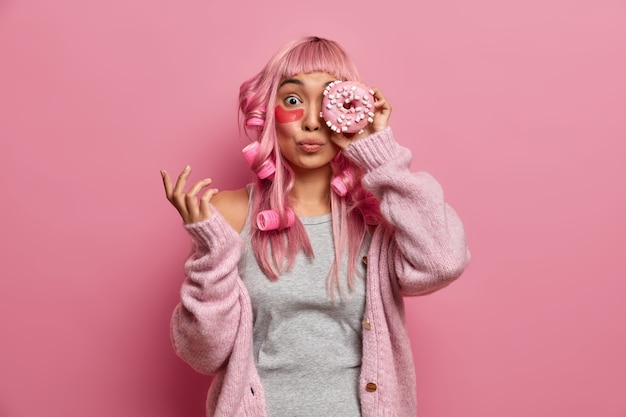Divertida mujer asiática de cabello rosado tiene rodillos en la cabeza, cubre los ojos con una deliciosa rosquilla dulce, usa parches de colágeno para reducir las arrugas