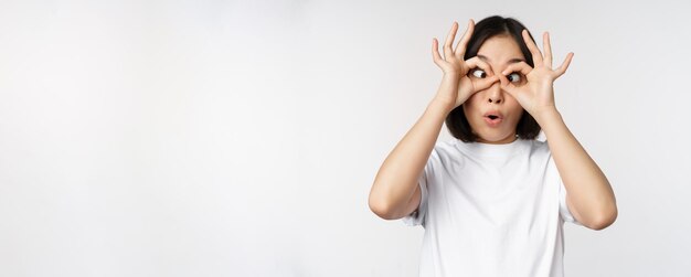 Divertida mujer asiática joven coreana haciendo ojos gafas gesto mirando feliz a la cámara de pie sobre fondo blanco.