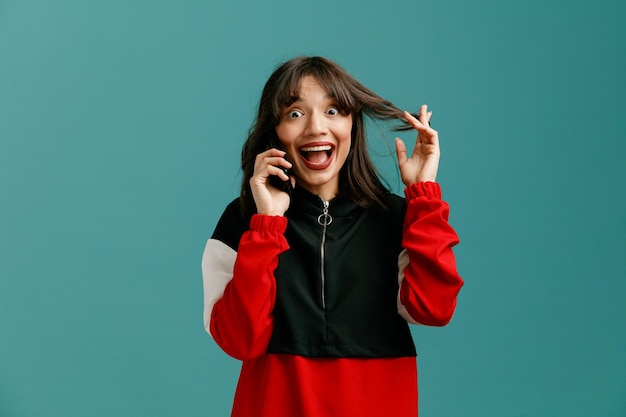 Divertida mujer caucásica joven agarrando el pelo mirando a la cámara mientras habla por teléfono aislado sobre fondo azul.