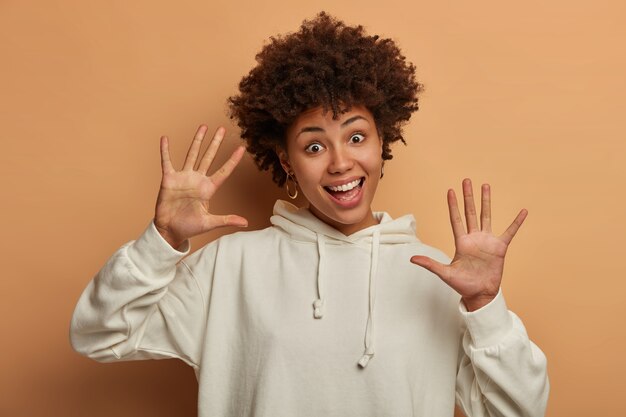 Divertida mujer étnica de piel oscura tiene el pelo rizado, se mueve con alegría, mantiene las palmas hacia la cámara, viste una sudadera blanca informal