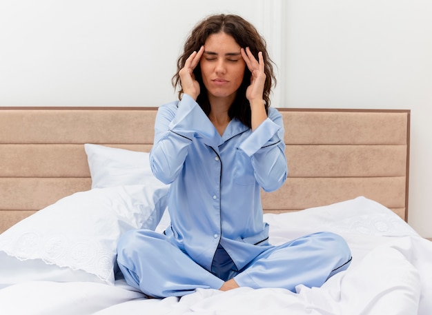 Divertida mujer hermosa joven en pijama azul sentada en la cama tocando las sienes mirando mal con dolor de cabeza en el interior del dormitorio