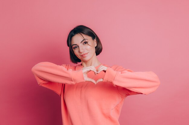 Divertida mujer joven de 20 años en ropa casual aislada en el retrato de estudio de fondo rosa. Concepto de amor. Muestra el corazón con las manos.