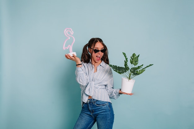 Divertida mujer joven expresiva con cabello oscuro con camisa azul y jeans posando con una sonrisa maravillosa con flamenco rosado y maceta contra la pared azul