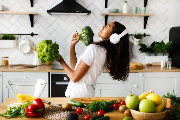 Divertida mujer mulata con grandes auriculares inalámbricos baila con hojas de ensalada y brócoli en la cocina moderna junto a la mesa llena de frutas y verduras