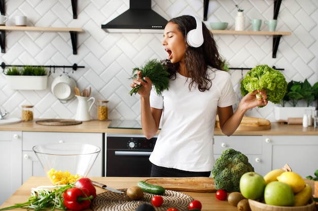 Divertida mujer mulata con grandes auriculares inalámbricos está cantando en un micrófono imaginario verde en la cocina moderna junto a la mesa llena de verduras y frutas