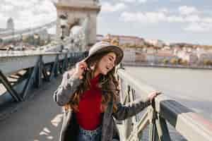 Foto gratuita divertida mujer de pelo largo con sombrero posando con los ojos cerrados durante la sesión de fotos en el puente en un día soleado