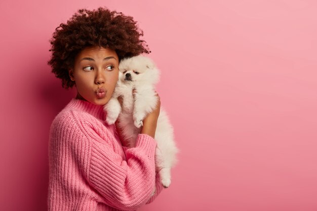 La divertida mujer de pelo rizado mantiene los labios doblados, disfruta del tiempo libre con un lindo cachorro de raza miniatura, mantiene al perro Spitz cerca de la cara