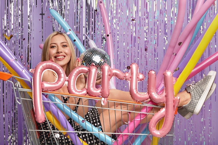 Foto gratuito divertida mujer rubia posa en carrito de compras con coloridos globos de modelado y bola de discoteca