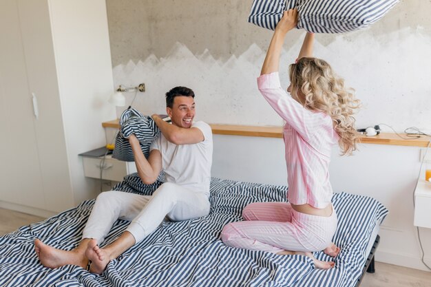 Divertida pareja joven divirtiéndose en la cama por la mañana, peleando con almohadas, jugando, sonriendo feliz