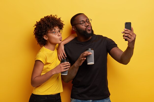 Divertida pareja de piel oscura hace pucheros con los labios a la cámara del teléfono celular, hace un retrato selfie, bebe café para ir de vasos desechables, usa camisetas negras y amarillas