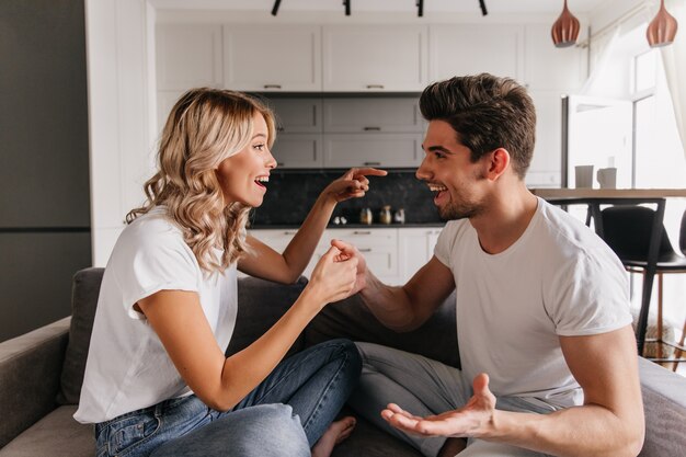 Divertida pareja sentada en el sofá, se mira y juega al juego de pulgar. Chica apuntando al chico y tratando de desviarlo.