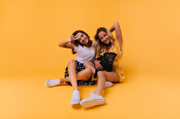 Divertidas chicas bronceadas posando en el suelo con perro. Retrato de impresionantes hermanas blancas aisladas en amarillo con bulldog francés.