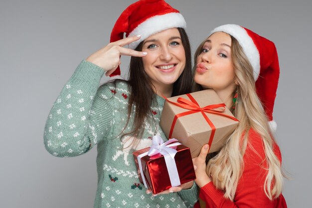 Divertidas mujeres con sombreros navideños rojos y blancos tiene regalos el uno para el otro y posa para la cámara