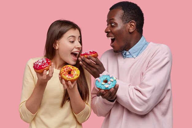 Divertido hombre y mujer joven de raza mixta prueban deliciosas donas, como postre dulce, pastelería, párate cerca, aislado sobre el espacio rosa