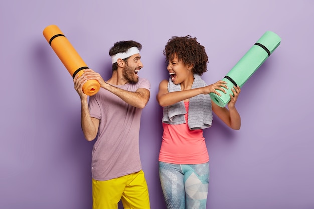 Divertido hombre y mujer multiétnica se divierten en el gimnasio, pelean con karemats enrollados