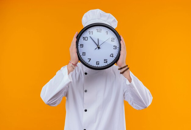Un divertido joven chef barbudo en uniforme blanco sosteniendo el reloj de pared frente a su cara en una pared naranja