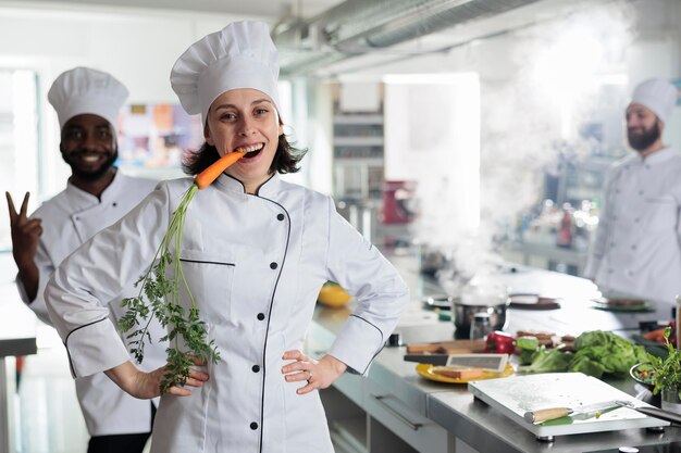 Divertido y juguetón jefe de cocina con zanahoria entre los dientes actuando tonto e infantil mientras posa para la cámara. Divertido experto en gastronomía con verduras en la boca sonriendo de corazón mientras está en la cocina del restaurante.