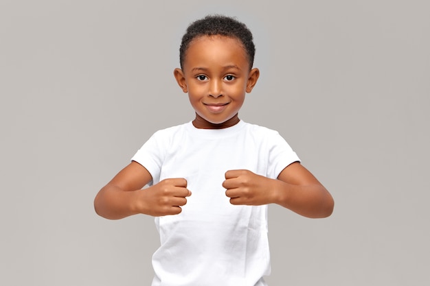 Foto gratuita divertido niño africano de diez años en camiseta blanca manteniendo los puños cerrados frente a él demostrando fuerza o sosteniendo objetos invisibles