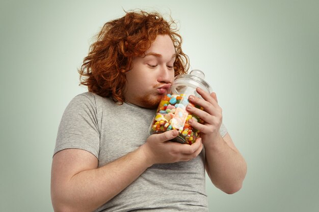 Divertido regordete joven varón caucásico con el pelo rizado de jengibre besando frasco de vidrio de dulces y mermeladas, abrazándolo suavemente al corazón. Concepto de obesidad, glotonería, alimentación, nutrición y estilo de vida poco saludable